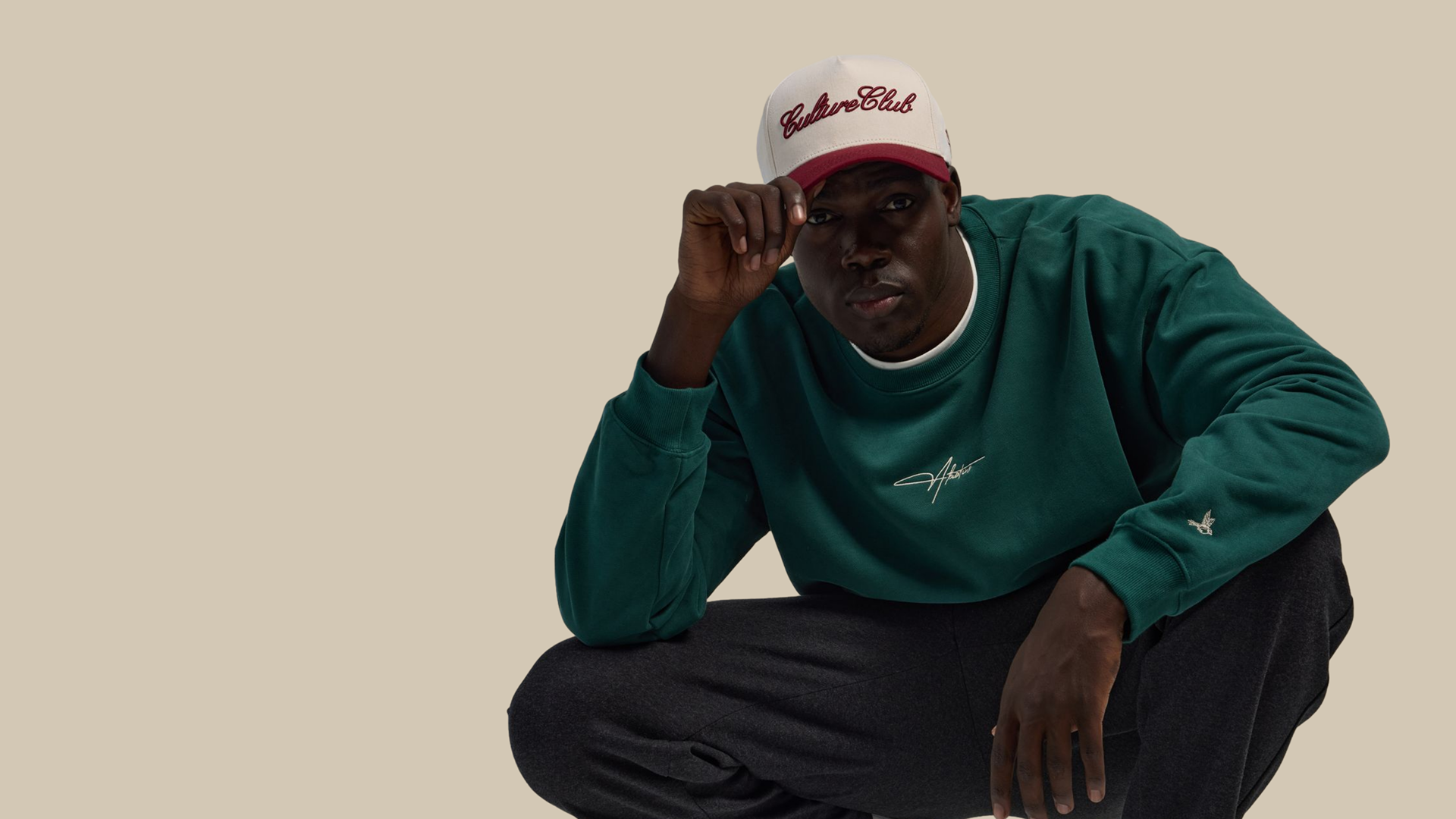 Person wearing a green sweatshirt and white cap with red brim on a beige background